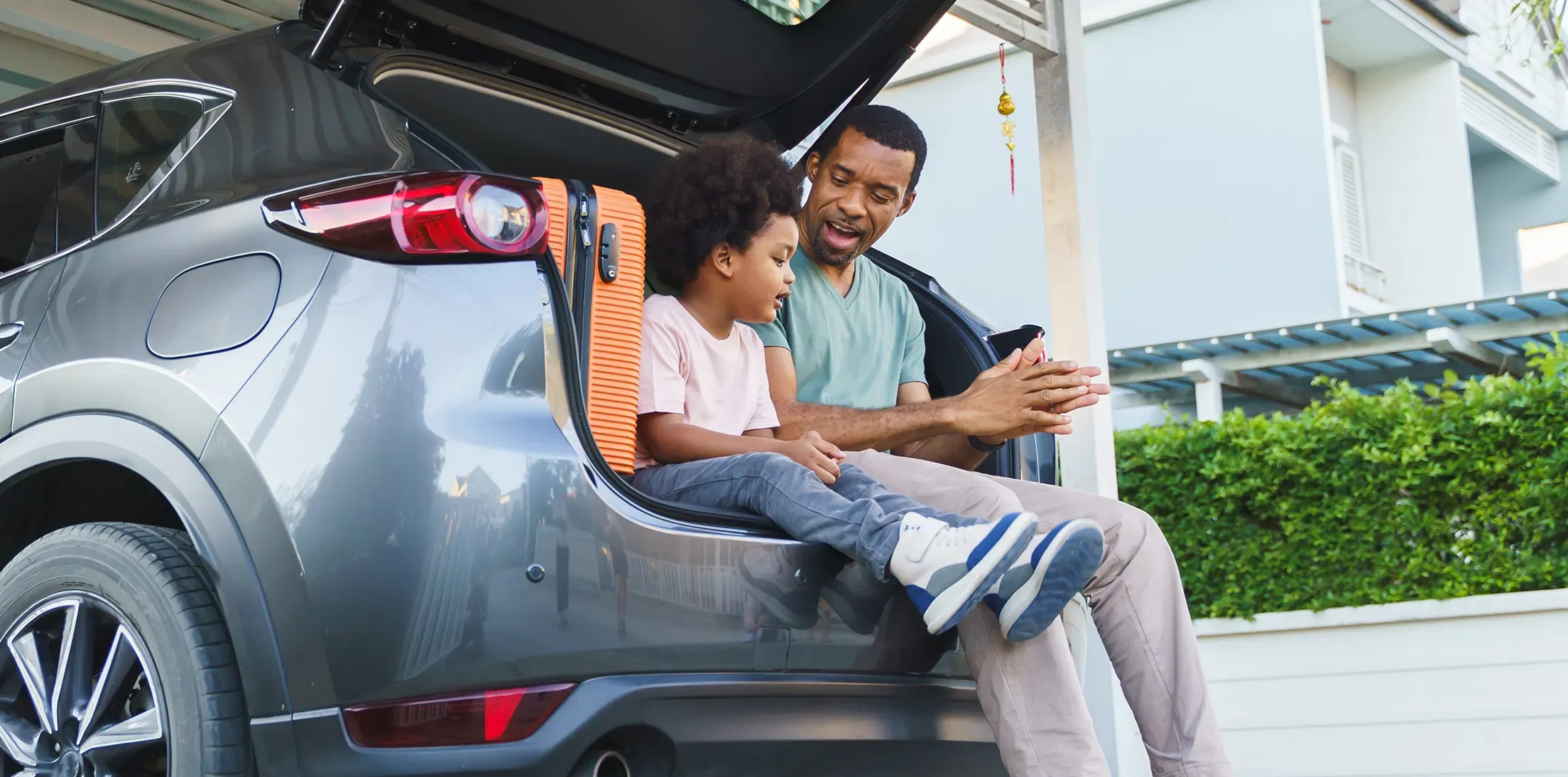 An adult and child sitting beside luggage in the open trunk of a car